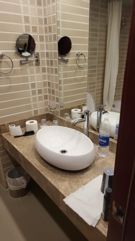 Ein sauberes und funktionales Badezimmer mit beige gefliesten Wänden, einem modernen Aufsatzwaschbecken auf einer Marmor-Waschtischplatte, einem großen Spiegel, zwei kleineren Vergrößerungsspiegeln und einer Wasserflasche.
