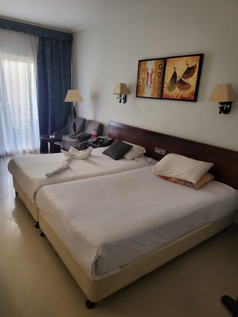 Ein helles Hotelzimmer mit zwei Einzelbetten, weißer Bettwäsche und einem dekorativ gefalteten Handtuch. An der Wand hängen zwei moderne Kunstbilder und Wandlampen. Im Hintergrund ist ein Sofa und der Zugang zu einem Balkon zu sehen.
