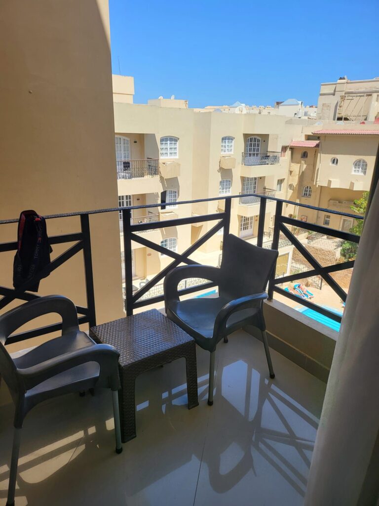 Ein sonniger Hotelbalkon mit zwei grauen Plastikstühlen und einem kleinen Tisch. Über das Geländer blickt man auf die umliegenden beigefarbenen Hotelgebäude und einen Poolbereich, in dem eine Person im Wasser entspannt.