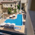 Eine Vogelperspektive auf einen Hotelpool in L-Form mit blauen Fliesen, umgeben von Liegestühlen mit weißen Sonnenschirmen. Die Poolanlage ist eingebettet zwischen mehrstöckigen, beigefarbenen Hotelgebäuden mit Balkonen, unter strahlend blauem Himmel.