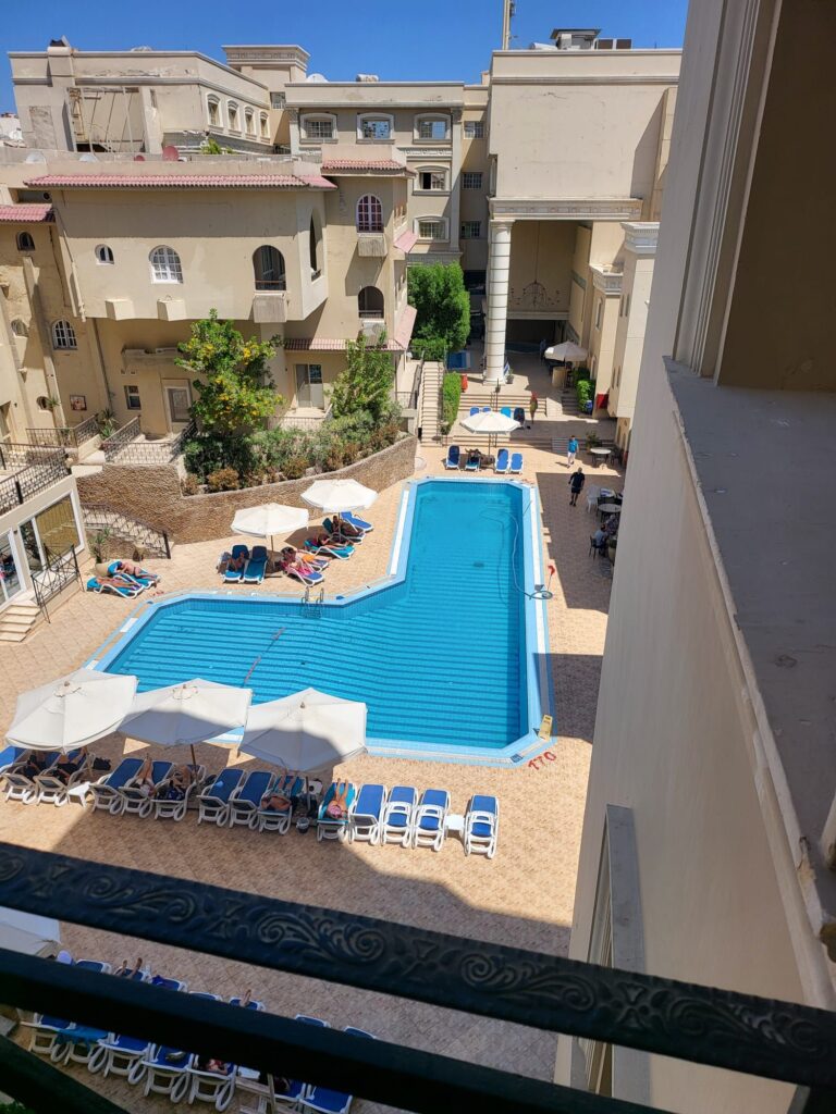 Eine Vogelperspektive auf einen Hotelpool in L-Form mit blauen Fliesen, umgeben von Liegestühlen mit weißen Sonnenschirmen. Die Poolanlage ist eingebettet zwischen mehrstöckigen, beigefarbenen Hotelgebäuden mit Balkonen, unter strahlend blauem Himmel.